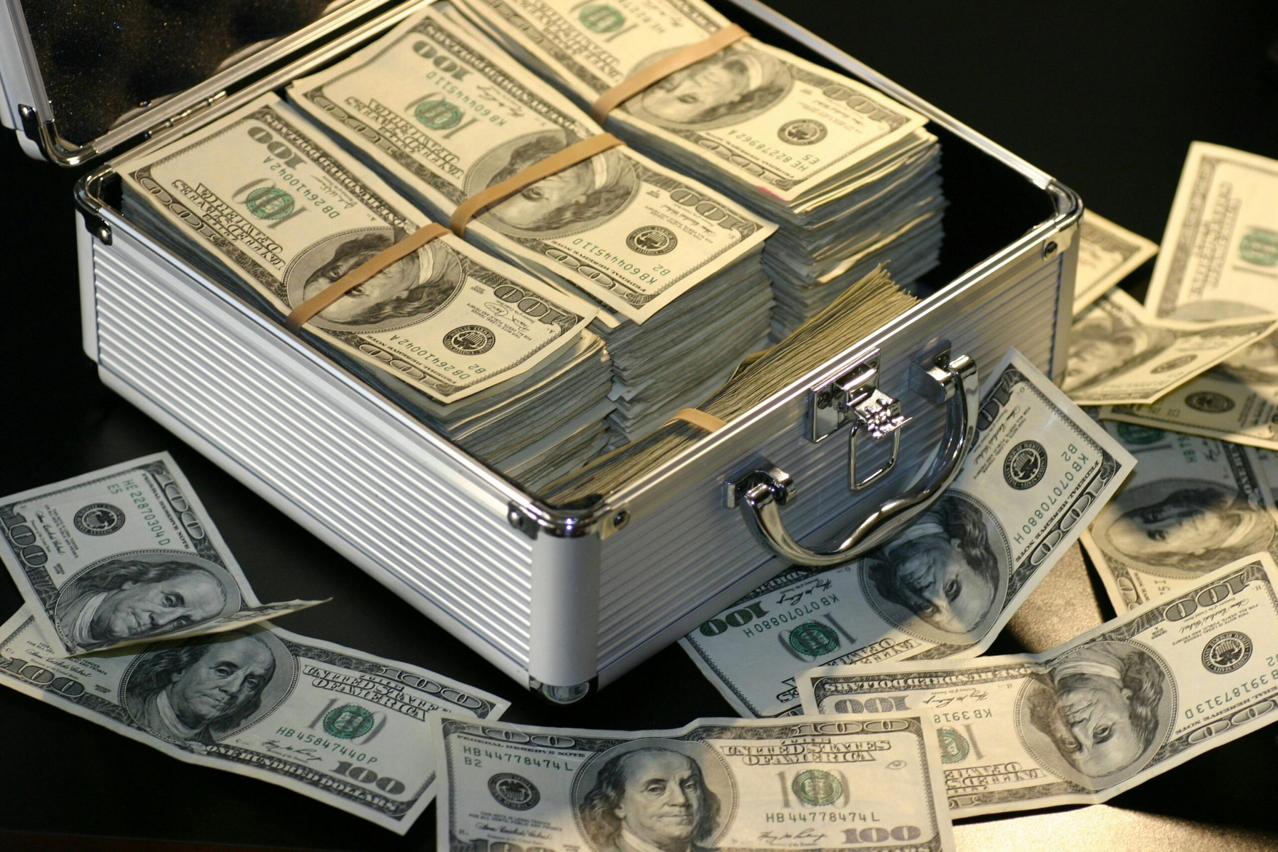 Stacks of $100 bills bound with bands inside a metal briefcase, money overflowing around it