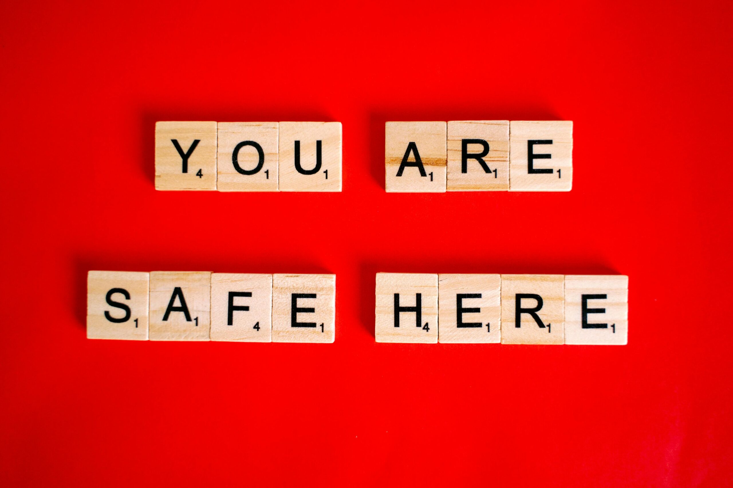 Scrabble tiles spell 'YOU ARE SAFE HERE' on a red surface, conveying a reassuring message.