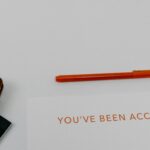 White page with the orange words "YOU'VE BEEN ACCEPTED"; an orange pen lies above, with a brown notebook and paper clips resting to the left, on a clean white surface.