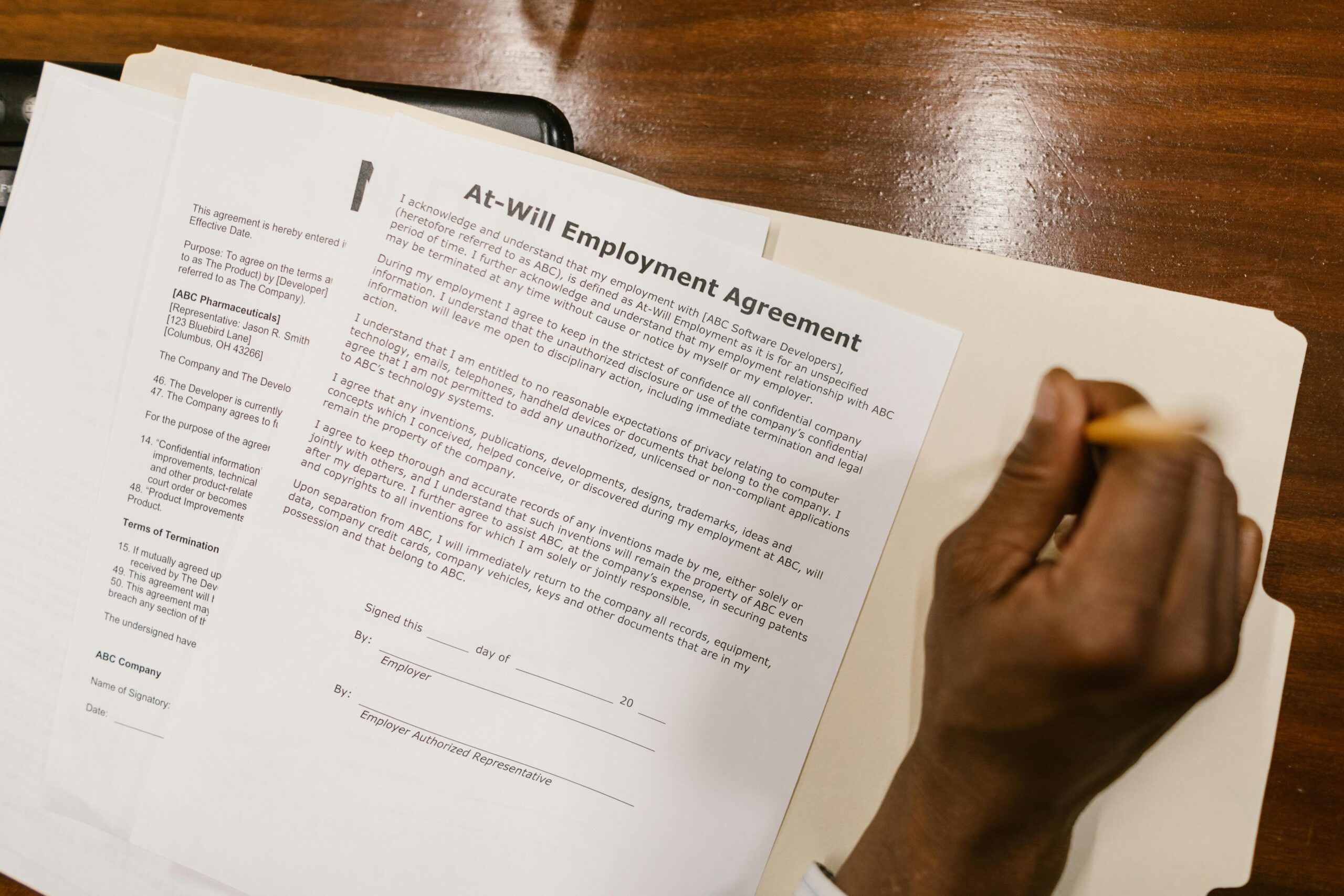 Hand signing an At-Will Employment Agreement on a wooden desk.