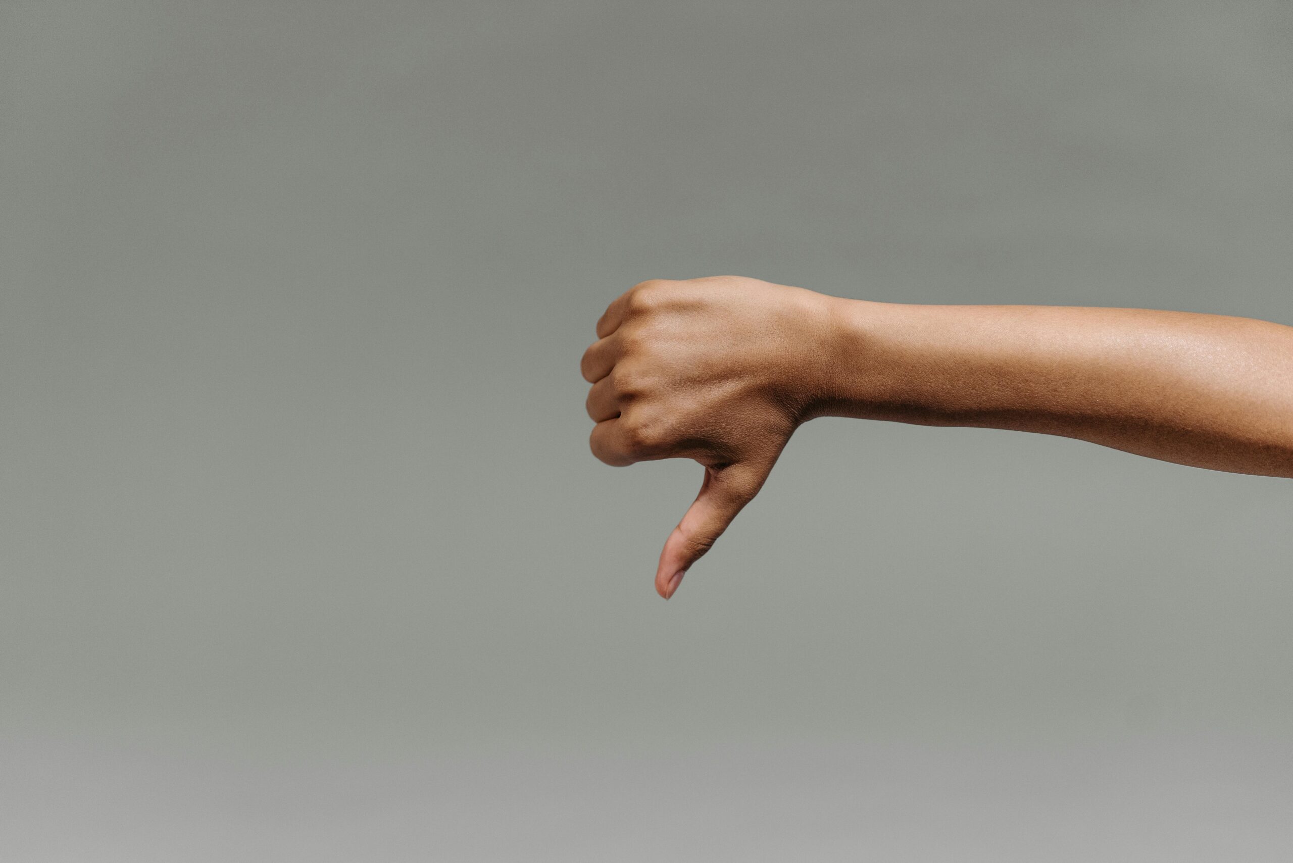 Arm extended to the right with a hand showing a thumbs-down gesture against a gray background, conveying disapproval